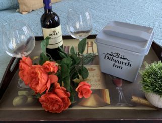 A tray holds two glasses and a bottle of wine plus fresh cut roses resting on a plush and colorful bed.