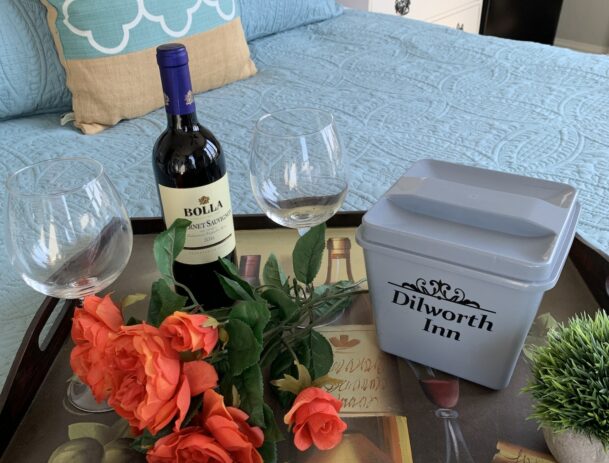 A tray holds two glasses and a bottle of wine plus fresh cut roses resting on a plush and colorful bed.
